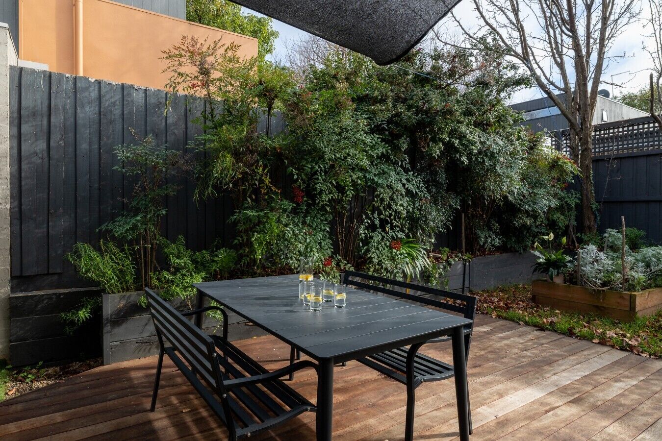 A patio with a table and benches, surrounded by dense foliage and a wooden fence.
