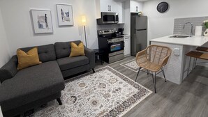 A modern living room with a sofa, a dining area with a chair, a kitchen with appliances, and a clock on the wall. - Modern Rangeview Stay| Near YMCA & Cineplex Cinema (Calgary)