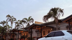 A white car parked in front of a building with a covered porch, palm trees, and a clear sky. - Lying Flat Hostel (Minxiong)