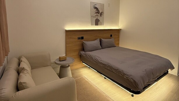A modern bedroom with a bed, a sofa, a small table, and a wall-mounted light fixture. - Lying Flat Hostel (Minxiong)