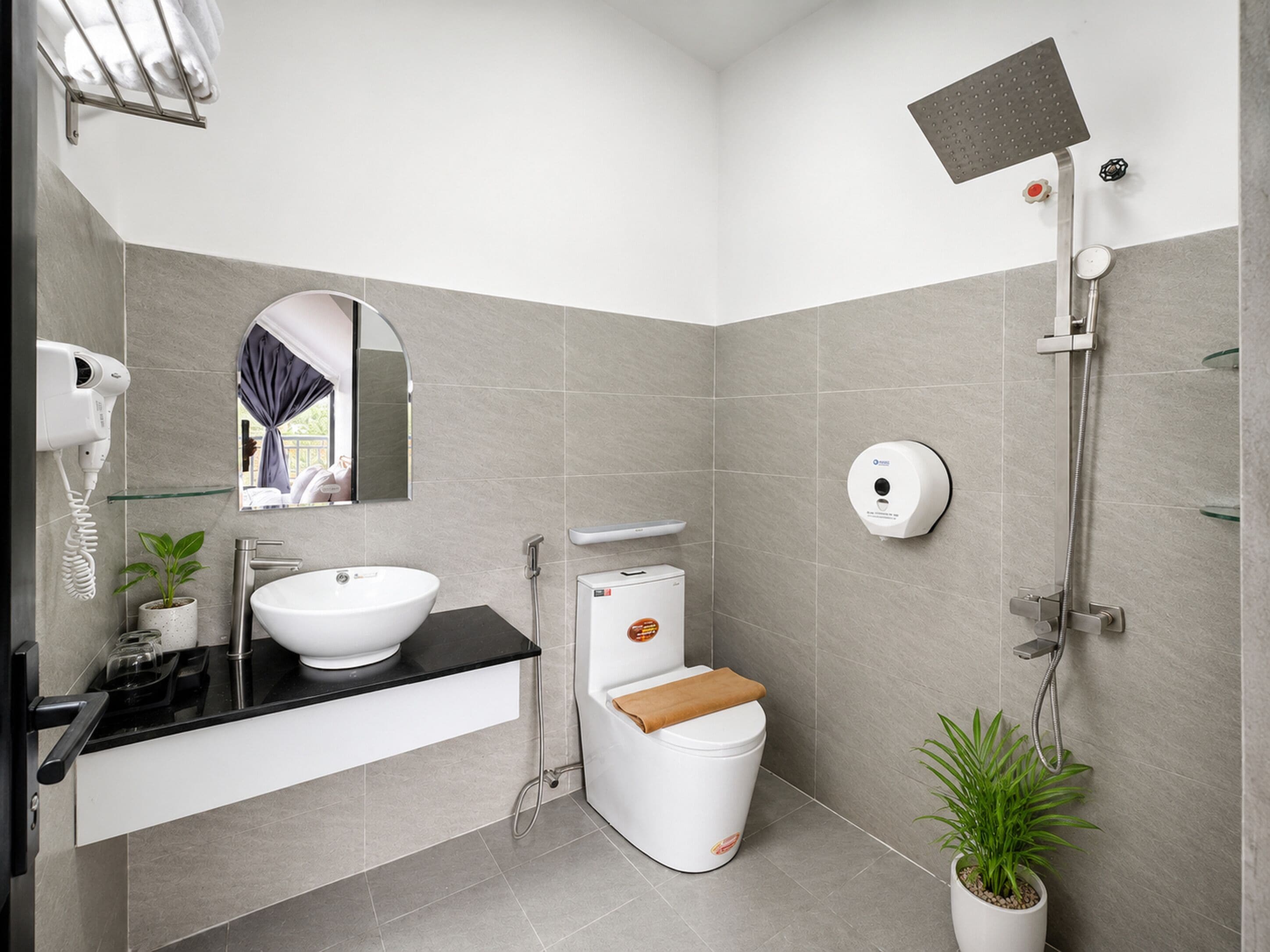 Une salle de bain moderne comprenant un lavabo, un miroir, un porte-serviettes, des toilettes, une douche et deux plantes en pot.