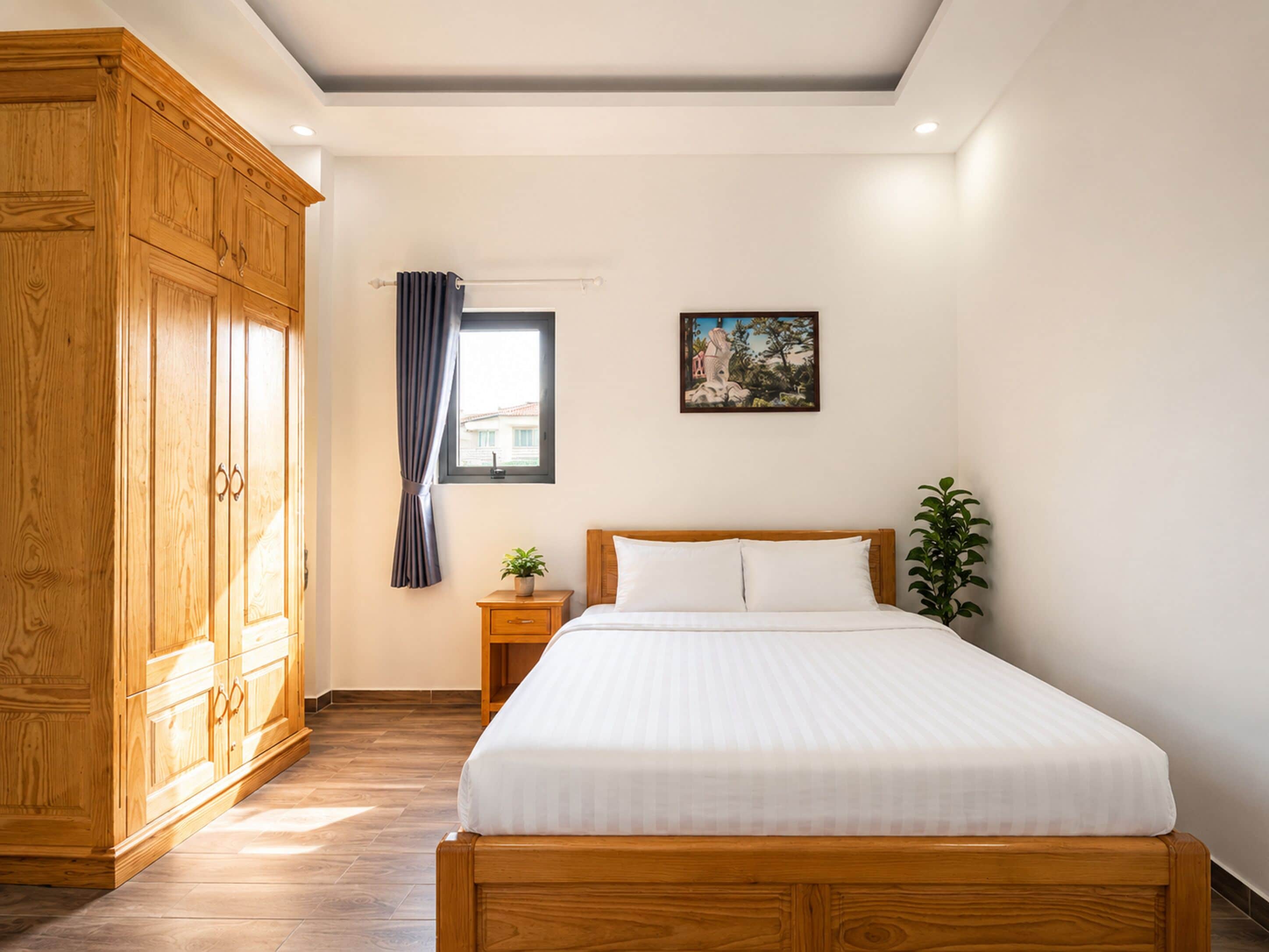 A bedroom with a large wooden wardrobe, a bed with white bedding, a small bedside table with a plant, and a window with curtains.