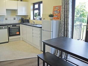 Cocina compacta con lavavajillas, horno y refrigerador, una mesa de comedor con banco, y un ventanal con vista a vegetación.