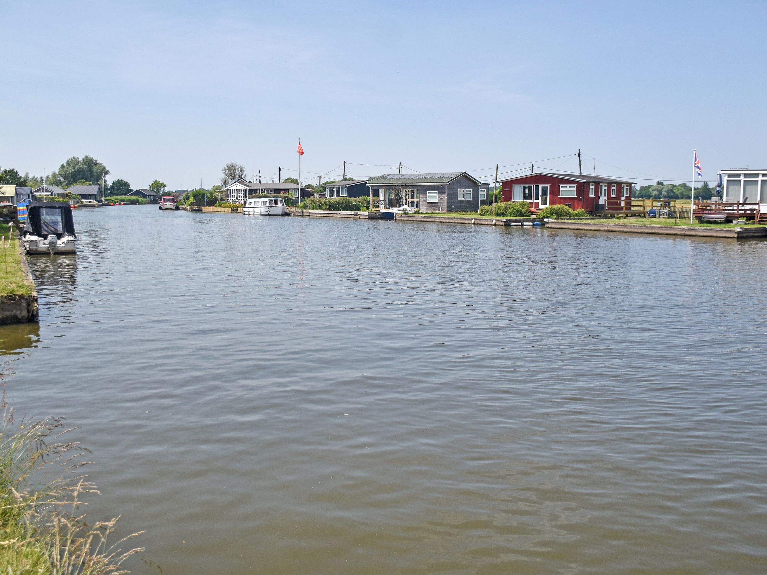 A calm river with boats docked on the left, houses along the riverbank, and flags on poles.