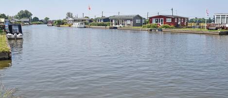 A calm river with boats docked on the left, houses along the riverbank, and flags on poles.
