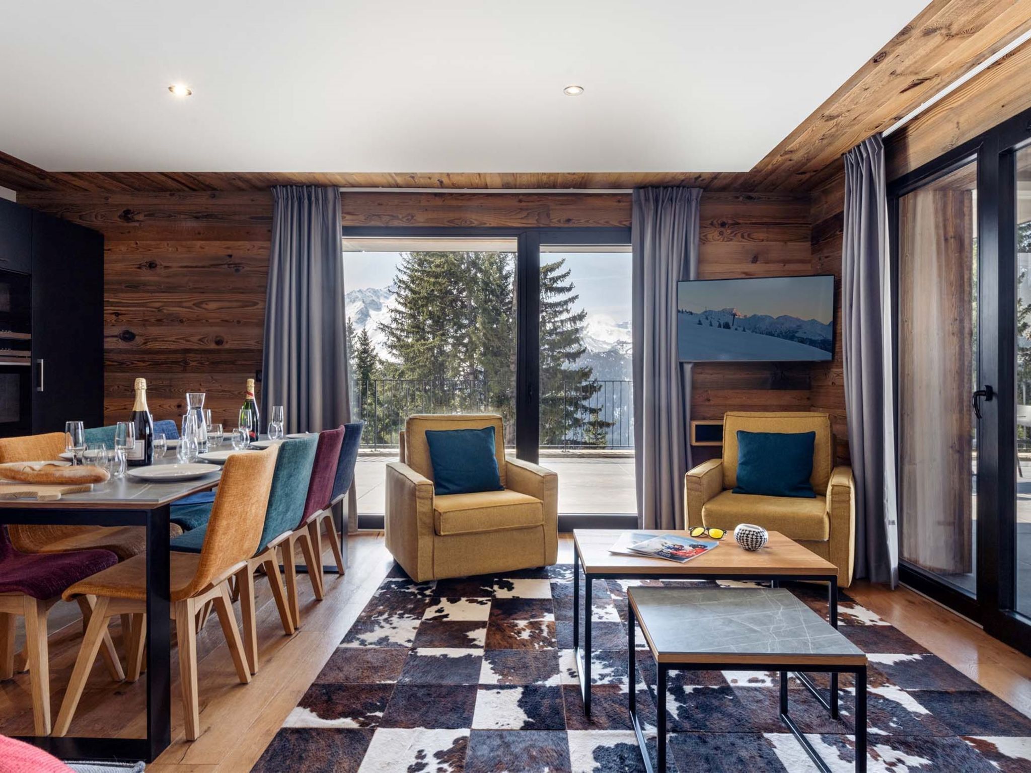 Un interior moderno con zona de comedor, sofás, una mesa de centro y un amplio ventanal con vistas a la montaña.