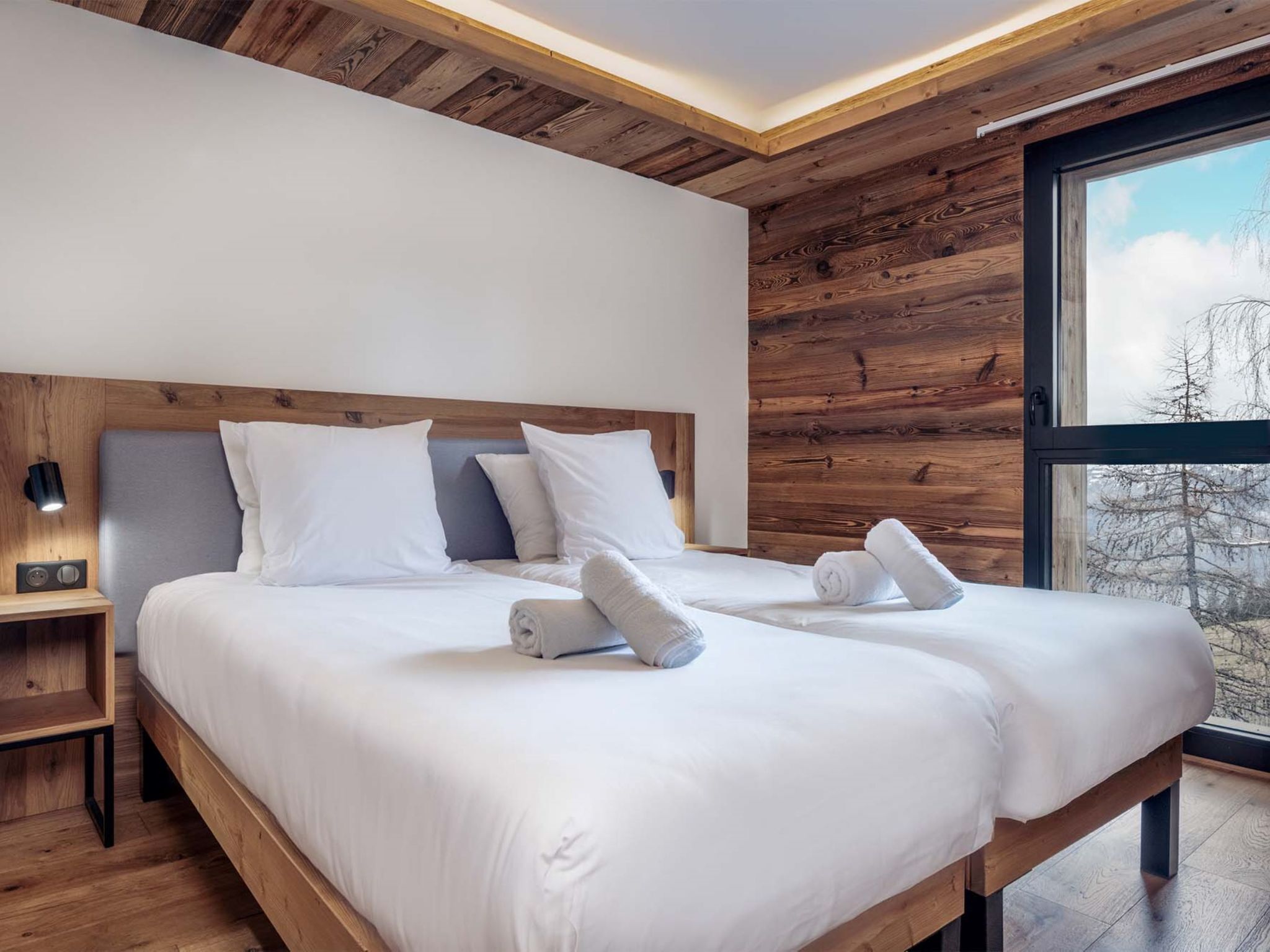 Una camera d'albergo moderna con soffitto in legno, due letti con lenzuola bianche, un comodino con una lampada e un'ampia finestra con vista sugli alberi.