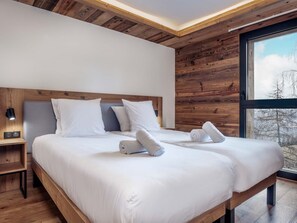 Ein modernes Hotelzimmer mit Holzboden, zwei Betten mit weißen Laken, einem Nachttisch mit Lampe und einem großen Fenster mit Blick auf Bäume.