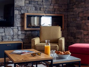 Ein gemütlicher Wohnbereich mit einer Steinwand, einem bequemen Sessel, einem Holztisch mit einer Zeitschrift, einer Flasche Orangensaft, einer Schüssel Croissants und einer Sonnenbrille.
