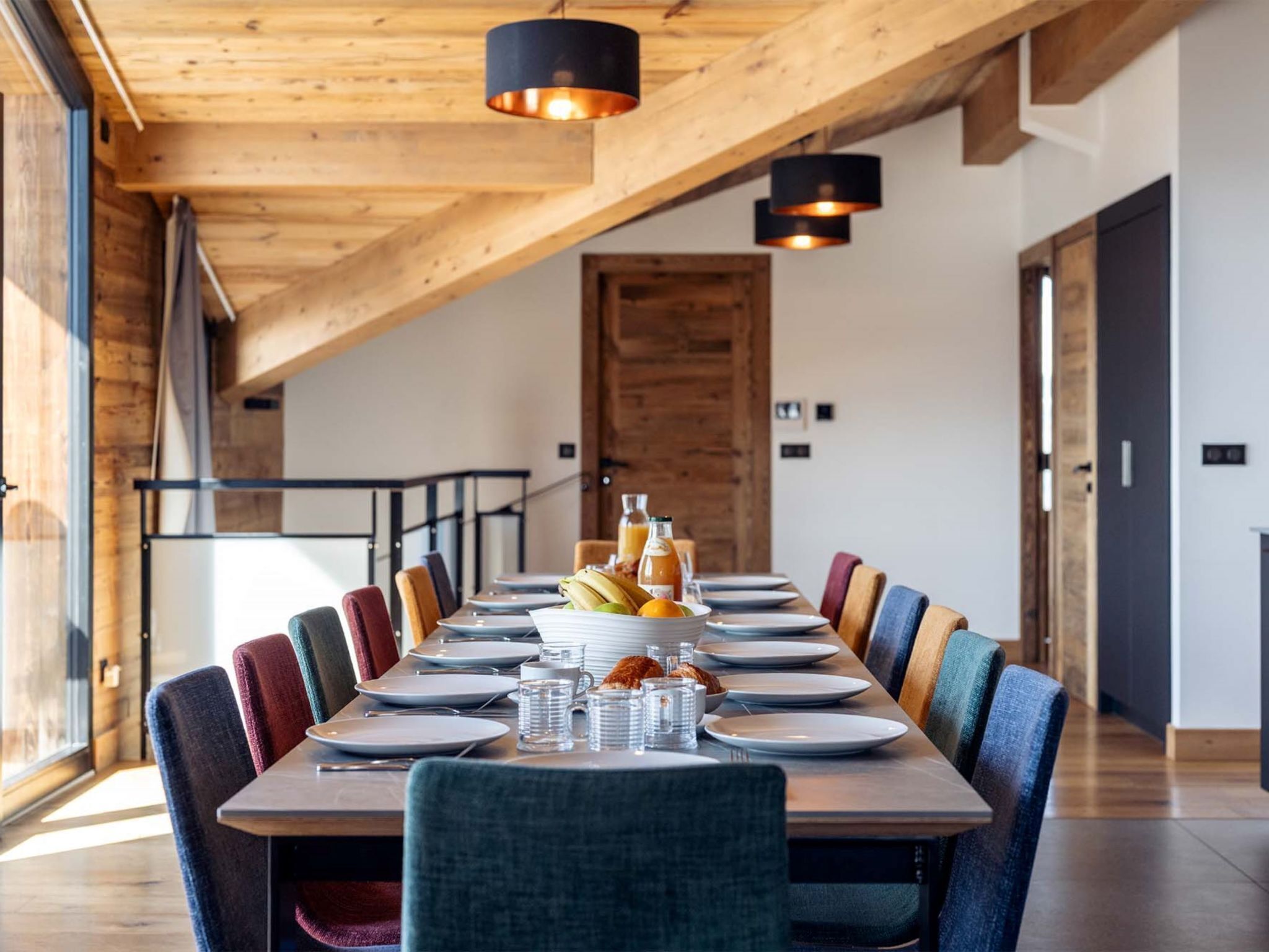 Un espace repas avec une longue table en bois, des chaises colorées et des luminaires suspendus.