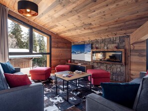Ein gemütliches Blockhaus-Interieur mit Holzwänden, einem Kamin aus Stein und einem großen Fenster mit Blick auf eine verschneite Landschaft.