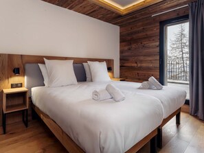 A modern bedroom with a wooden headboard, a large bed with white linens, two bedside tables with lamps, and a view of trees through a window. - Résidence l'Orée du Bois by Interhome (La Rosière)