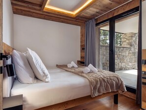 A bedroom with a wooden ceiling, a bed with white bedding, a stone wall outside, and a view of the outdoors. - Résidence l'Orée du Bois by Interhome (La Rosière)