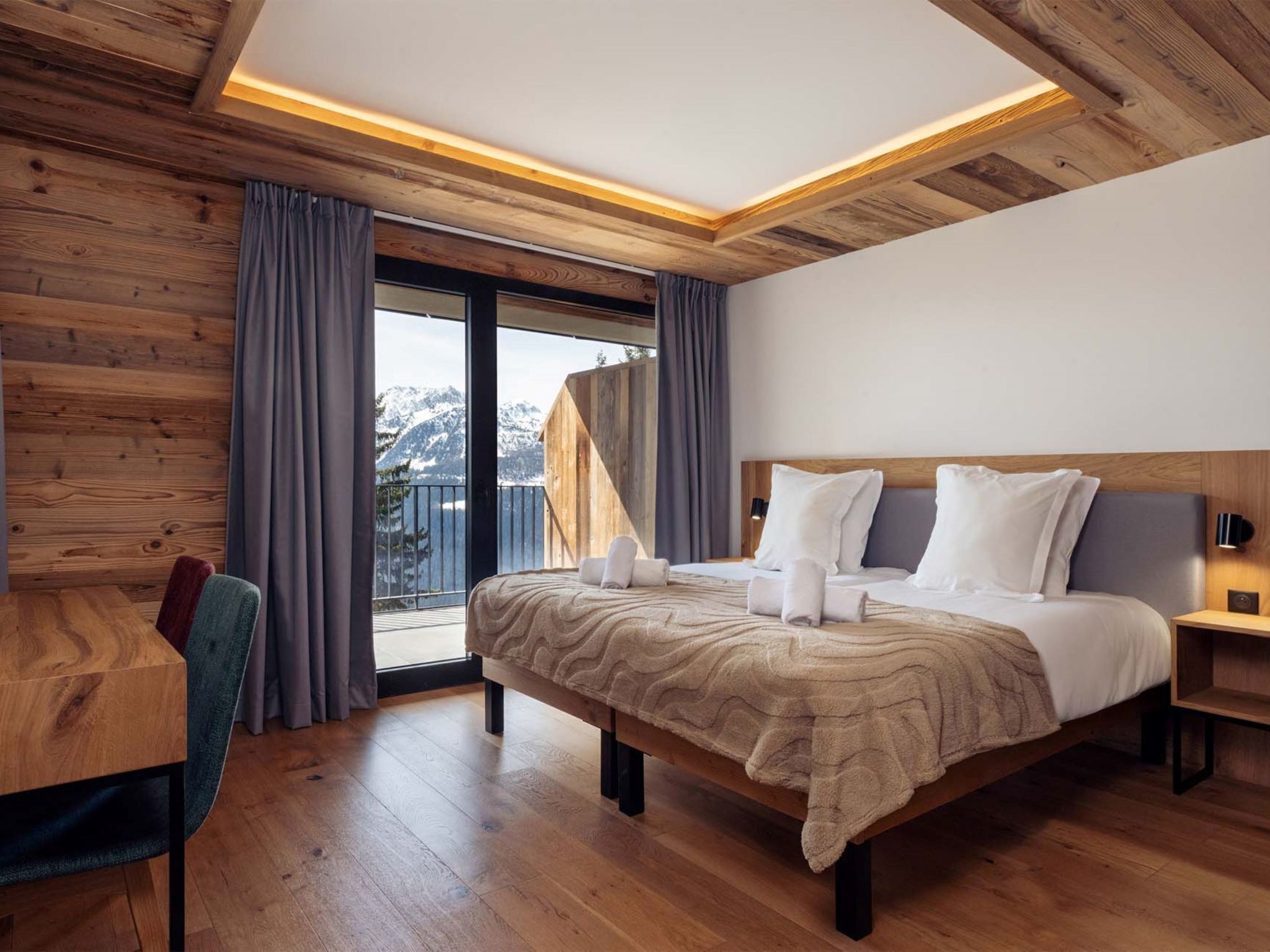 Una habitación de hotel moderna con techo de madera, una cama grande con ropa blanca, un escritorio de madera y vista a las montañas a través de la ventana.