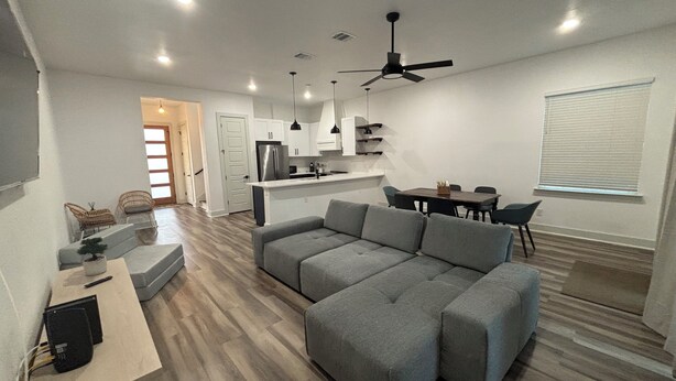 A modern living room with a sectional sofa, a dining table with chairs, a kitchen area, and a ceiling fan. - Newly Built Home 7 Min Drive River Walk & Downtown - 1222 (San Antonio)