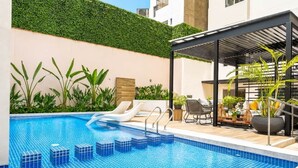 Una piscina con gradini, lettini e una zona relax coperta con piante.