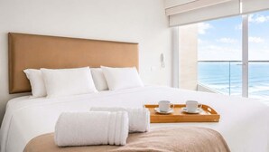 Una camera d'albergo con un letto grande, biancheria bianca, una testiera in legno, un vassoio di legno con una tazza e un piattino, e vista sull'oceano.