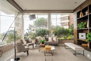 A modern living room with a sofa, chairs, a coffee table, and a bench. There is a floor lamp, a bookshelf with plants, and large windows offering a view of the outdoors. - Ocean View Luxury Apt | King Bed |Walk to Larcomar (Miraflores)