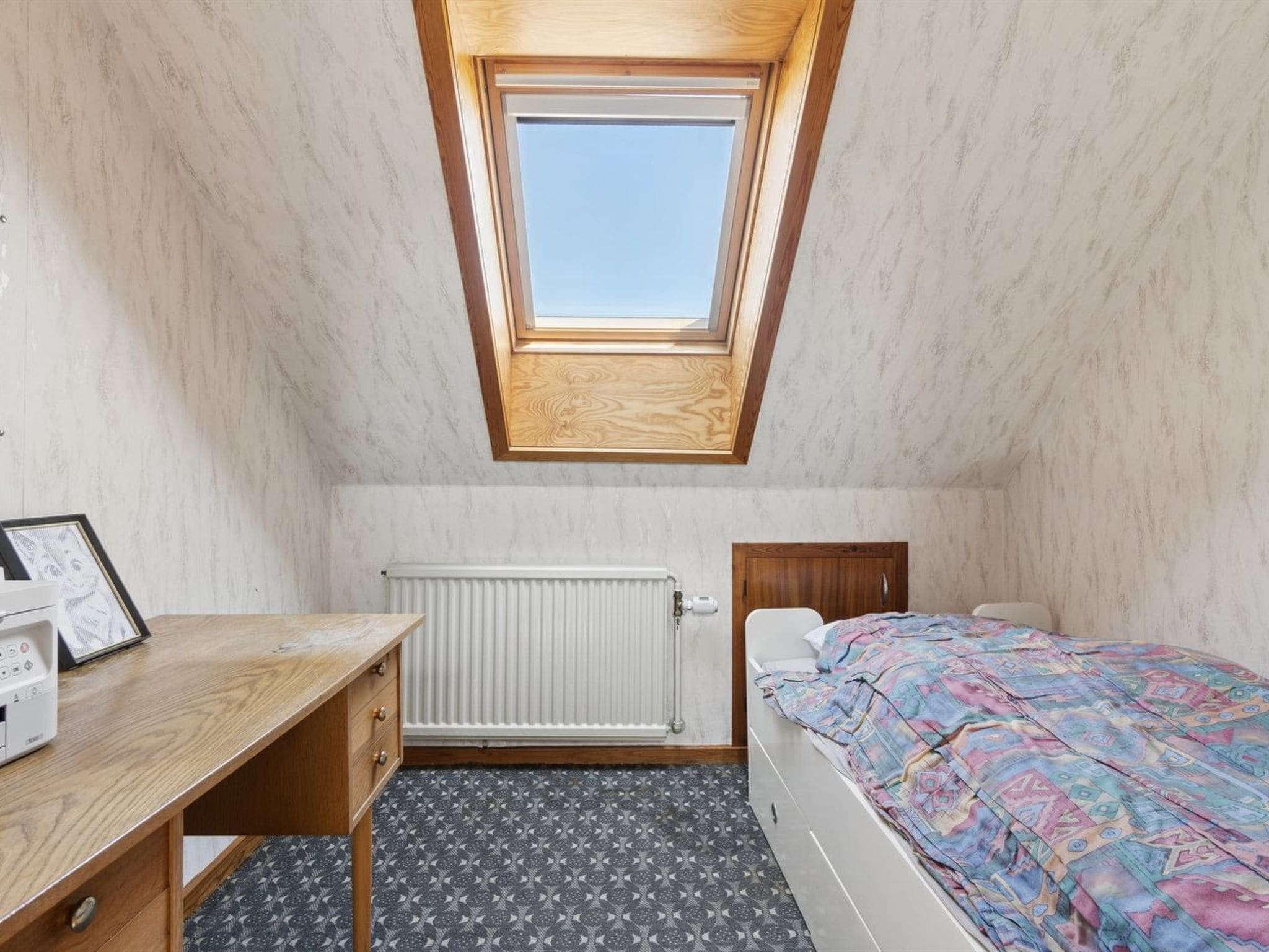 Une petite pièce bien éclairée, avec un plafond en pente, un bureau en bois, un lit recouvert d’une couverture colorée et un radiateur.