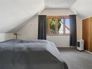 Ein Schlafzimmer mit schrägverdeckter Decke, einem Bett mit grauer Bettdecke, einem Fenster mit Vorhängen und einem Heizkörper.