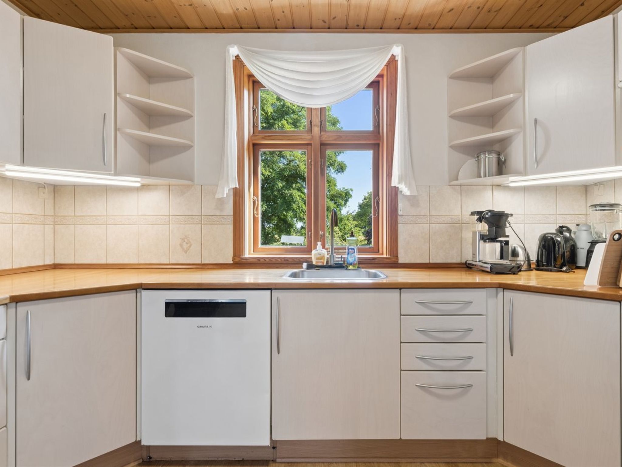 Cocina moderna con alacenas blancas, lavavajillas integrado, ventana con vista a zonas verdes y encimera con electrodomésticos.