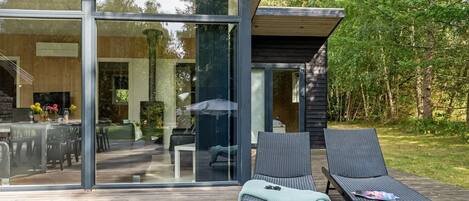 A modern outdoor deck with two lounge chairs and a dining area visible through large glass doors.