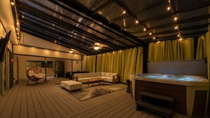 A cozy indoor space with a wooden deck, a sofa, a chair, a hot tub, and a ceiling with exposed beams and string lights.