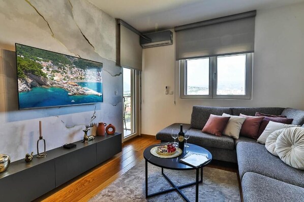 A living room with a large flat-screen TV, a grey sofa, a coffee table, and a view of a coastal town.