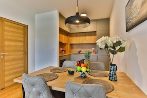 A dining area with a wooden table, grey upholstered chairs, a fruit bowl centerpiece, a hanging pendant light, and a vase with flowers.