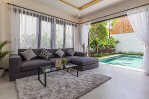 Un salon moderne avec un canapé gris, des coussins aux motifs noir et blanc, une table basse en verre et une vue sur une piscine.