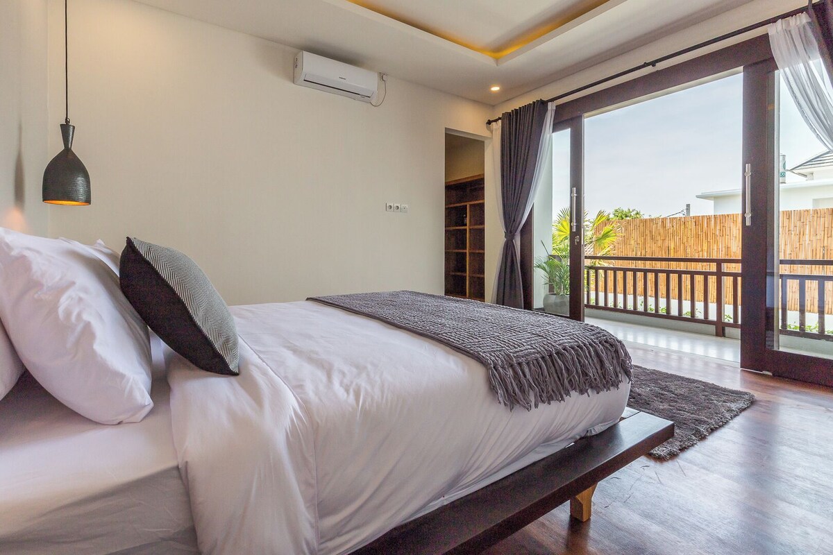 A bedroom with a bed, a bench, a hanging light fixture, and a view of the outside.