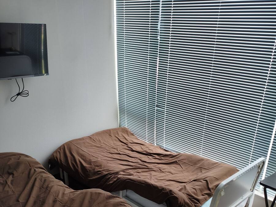 A room with two single beds, a television mounted on the wall, and a window with blinds.