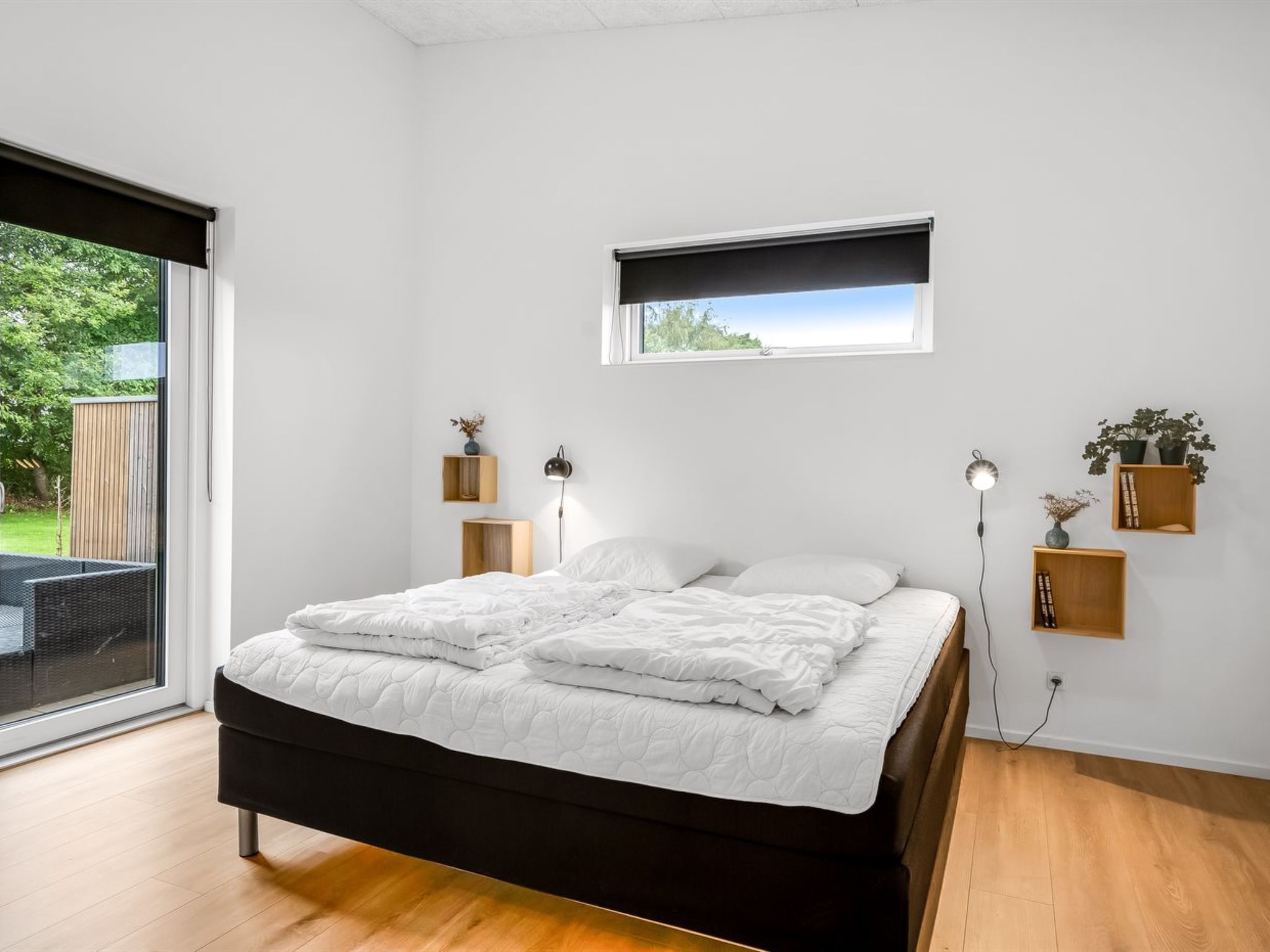 Een moderne slaapkamer met een groot bed, een raam met uitzicht, nachtkastjes met lampen en een klein plankje met planten.