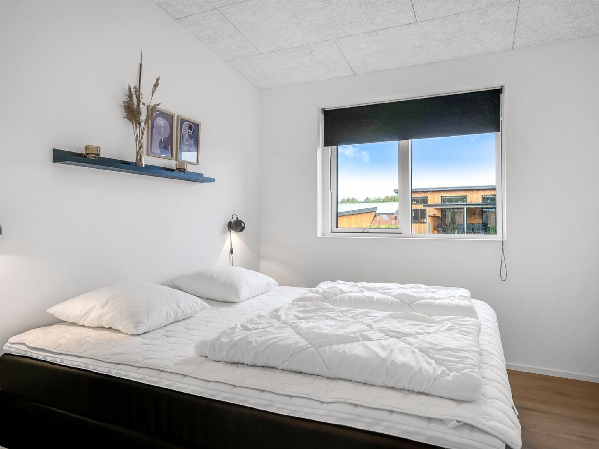 Ein Schlafzimmer mit einem Bett, einem Fenster mit Blick auf Gebäude, einem Regal mit Dekoration und einer Wandleuchte.