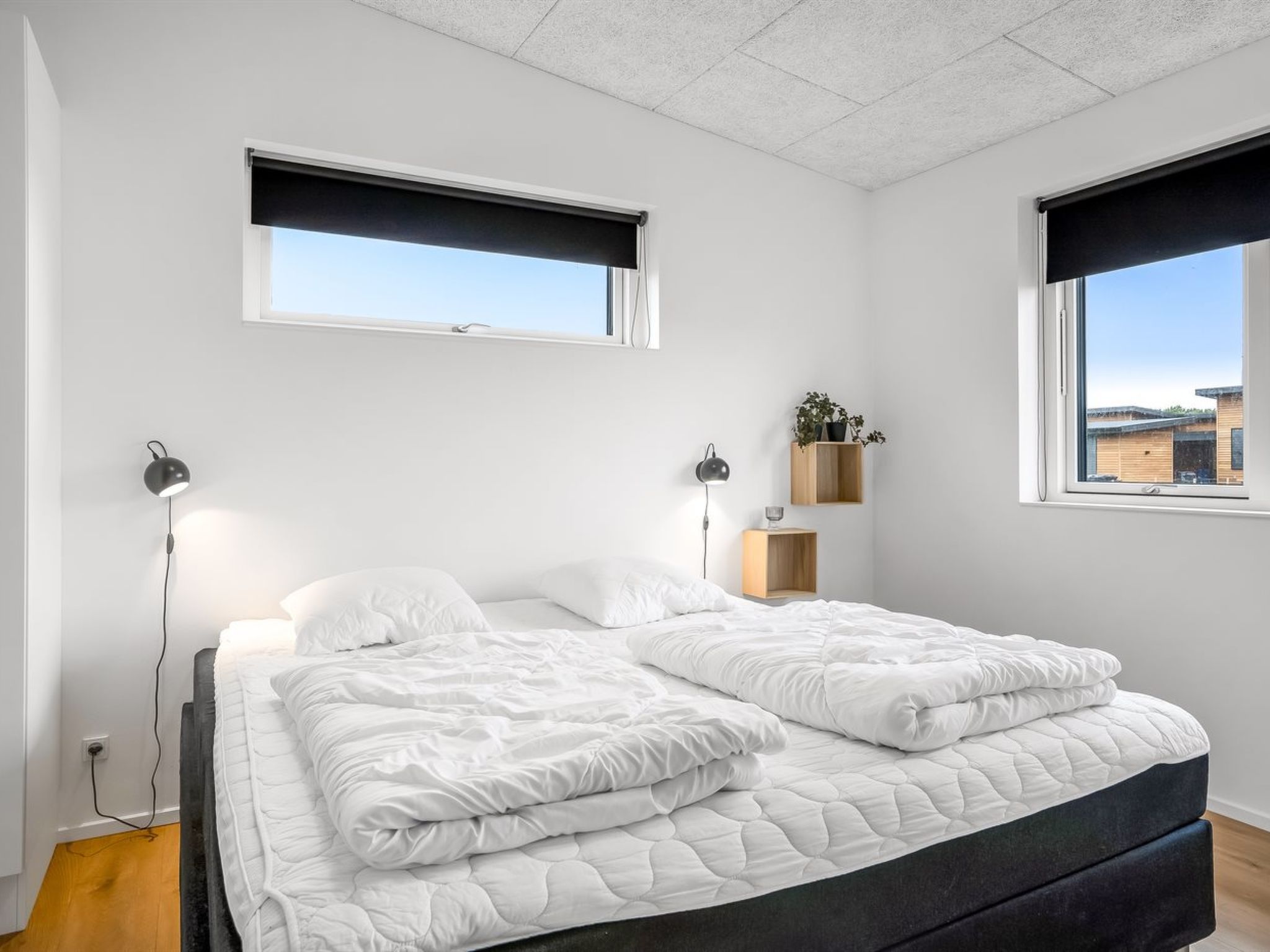 Un dormitorio con cama, dos lámparas de noche, un ventanal con vistas y una pequeña estantería con una planta.