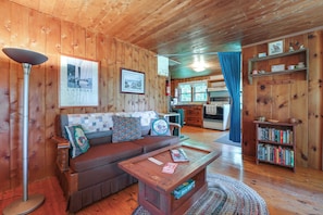 A cozy cabin interior with a wooden floor, a sofa with patterned cushions, a coffee table, a bookshelf, and a kitchen area visible through a doorway.