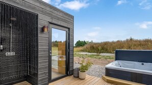 A modern outdoor hot tub area with a wooden deck, a glass door leading to a room, and a view of the surrounding landscape.