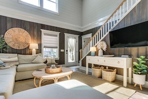 A living room with a sofa, a round wicker table, a TV, a staircase, and a large window. - Walk to Old Town Bluffton! Family Home w/ Balcony (Bluffton)