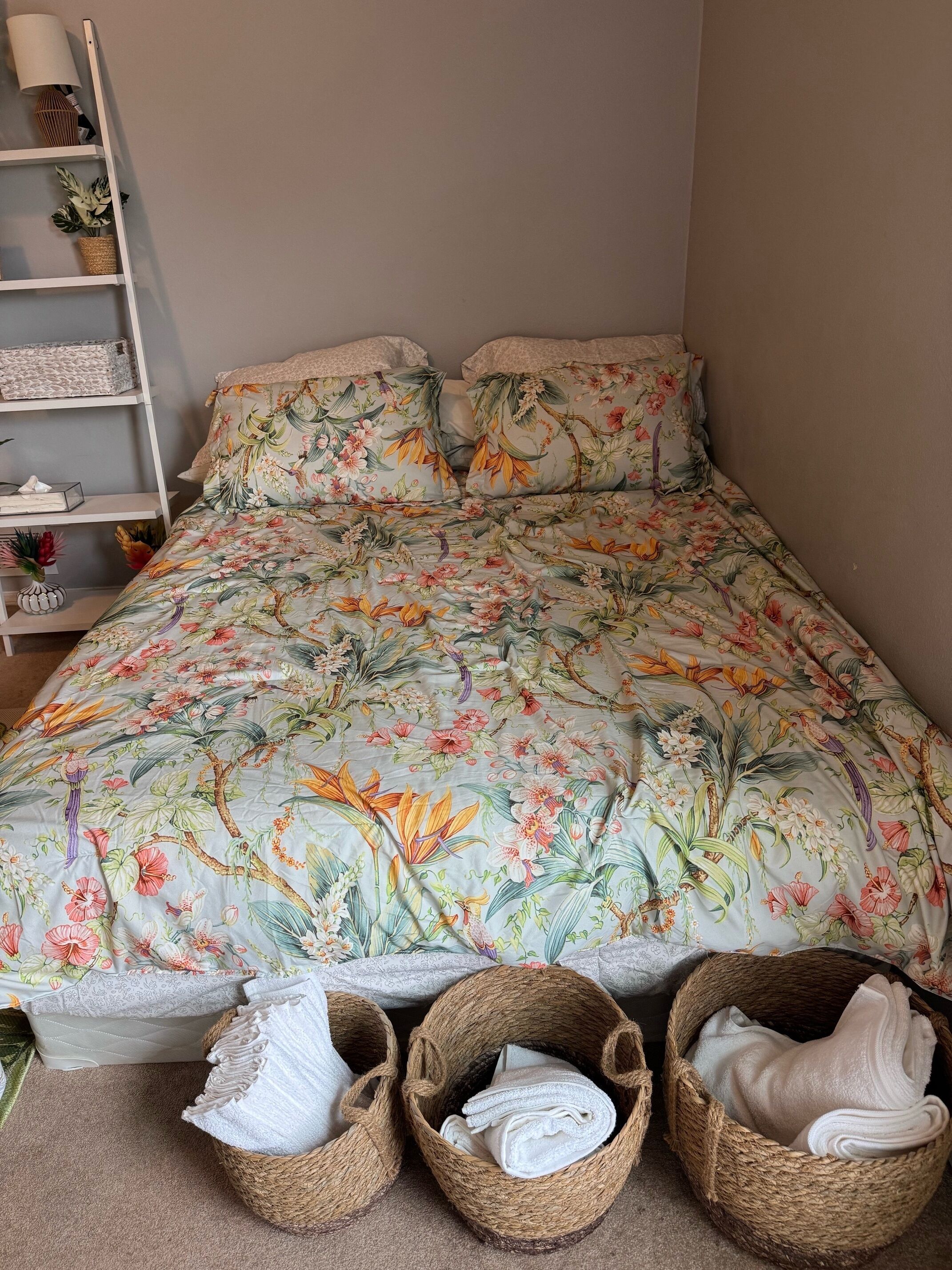 A bedroom with a floral-patterned bedspread, a bedside shelf with decorative items, and two woven baskets on the floor.