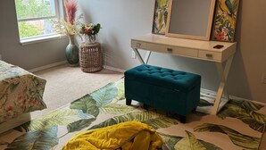 A bedroom with a bed, a desk with a mirror, a window, and a tropical-themed rug.