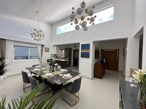 A modern dining area with a large table set for six, a chandelier, and a view of the sky through a window. - Modern Luxury Villa - Vista Cana  Private Pool- Golf - Beach Access (Punta Cana)