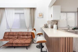 Ein modernes Wohnzimmer mit einer braunen Couch, einem Essbereich mit Stühlen, einer Küche mit weißer Arbeitsplatte und einem Fenster mit Vorhängen.
