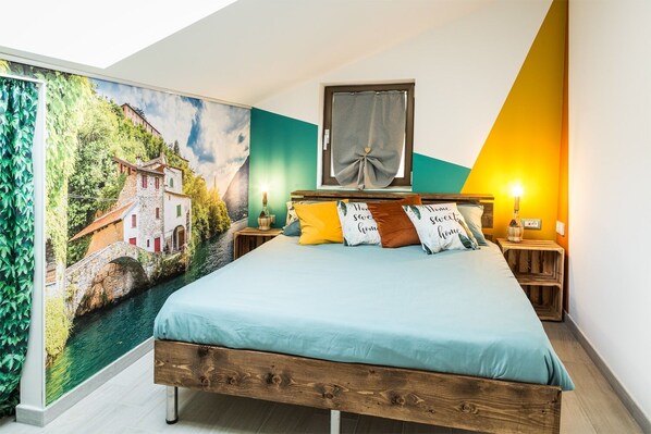 A bedroom with a bed, pillows, a nightstand, a lamp, a window with curtains, and a mural on the wall.