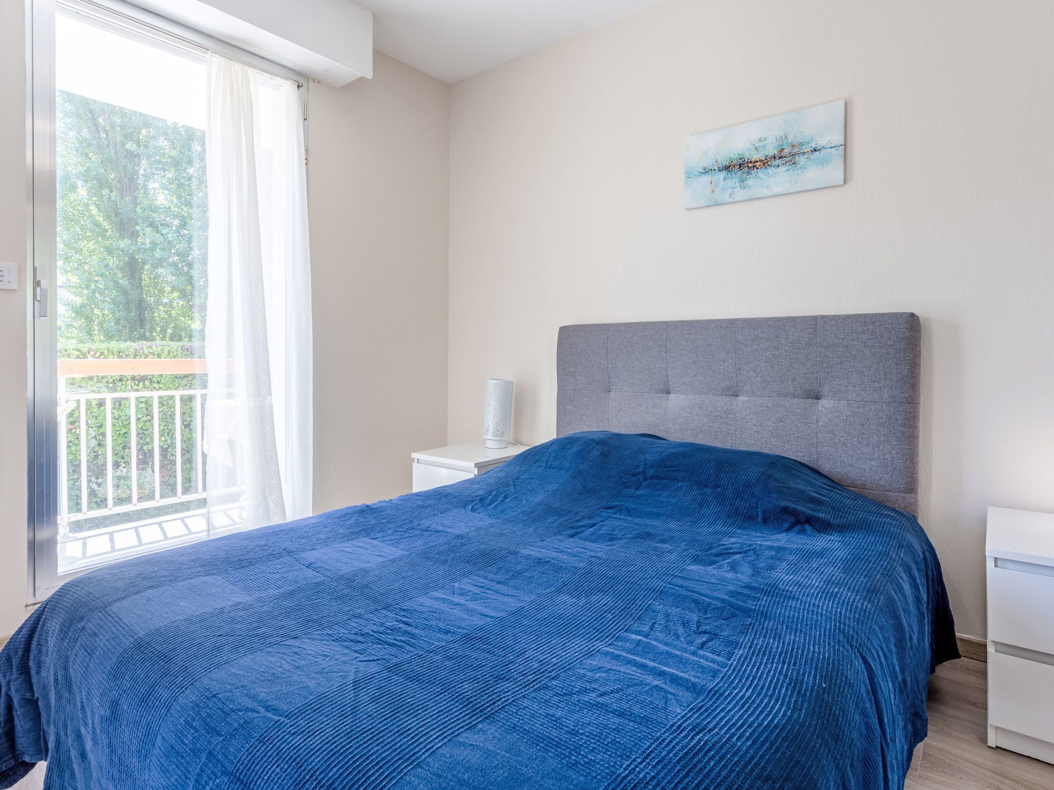 A bedroom with a bed, a nightstand, a lamp, a painting on the wall, and a balcony with greenery.