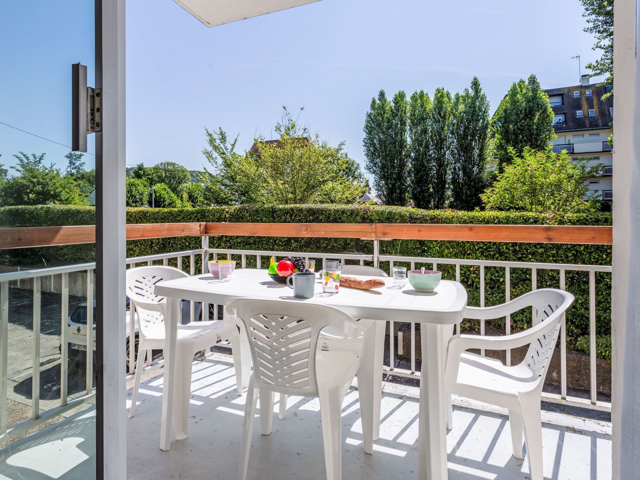 Un balcon avec une table et des chaises blanches, une clôture en bois et une vue sur les arbres et les bâtiments.