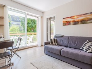 Una sala de estar moderna con un sofá gris, una mesa de comedor con sillas y un ventanal con vistas a zonas verdes y edificios.
