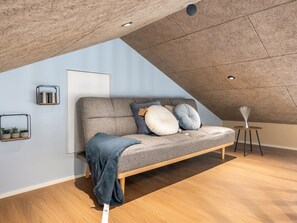 Ein Loft-Zimmer mit schrägem Dach, einem grauen Sofa mit Kissen, einem kleinen Beistelltisch und einer Wandablage mit Pflanzen.