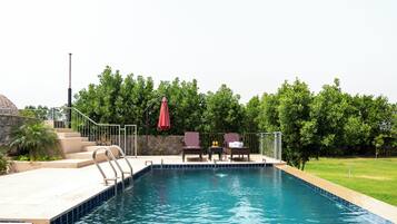 Una piscina con borde de piedra, una escalera metálica y una zona de estar con sillas y una mesa.