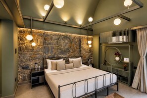 A bedroom with a stone wall, a bed with multiple pillows, bedside tables, a mirror, and a shelf with books and decorative items. - ELIVAAS Stone Age 6BHK Villa with Pool (Jalsu)