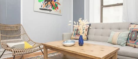 Un salón con una silla de ratán, un sofá con cojines estampados, una mesa de centro de madera y un cuadro abstracto y colorido en la pared.